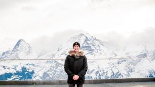 A young Asian man in a black coat with the Swiss Alps behind him