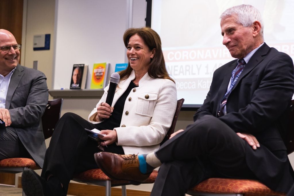 A woman in a white jacket laughs into a microphone. She is seated next to Dr. Anthony Fauci.