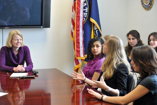 Students sit at a table with Lauren McFerran with a TV and the United States and United States Senate flags in the background