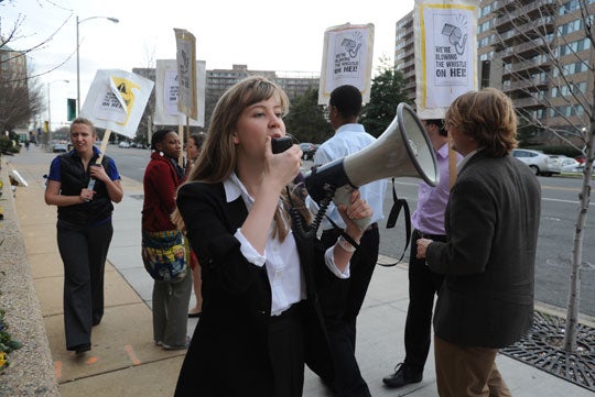 Julia Hubbell speaks into a megaphone while others march with picket signs behind her