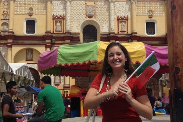 Lauren Barrison wears colorful clothes in Mexico, a yellow brick building and cafe umbrella are behind her