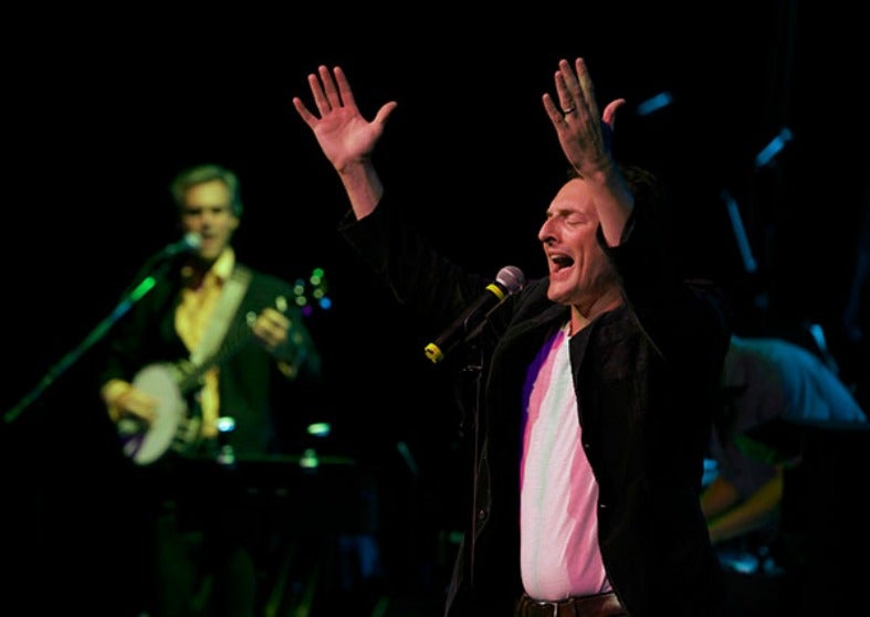 Rich Battista raises his hands above his head as he sings on stage at a rock concert, a guitar player stands behind him