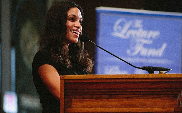 Rosario Dawson speaks behind a podium with a pop up banner for the Lecture Fund in the background