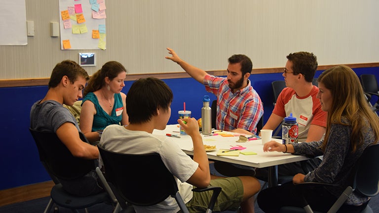 A student gestures at a poster board with multi-colored post-it notes as a group of students sitting at a table look on.