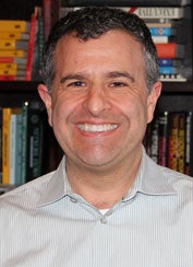 Peter Luger smiles for a headshot against a background of bookshelves. 