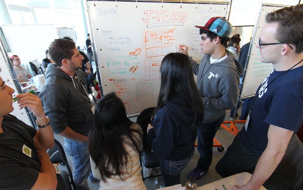 A group of students writes on a whiteboard in red and black; one member points at the board