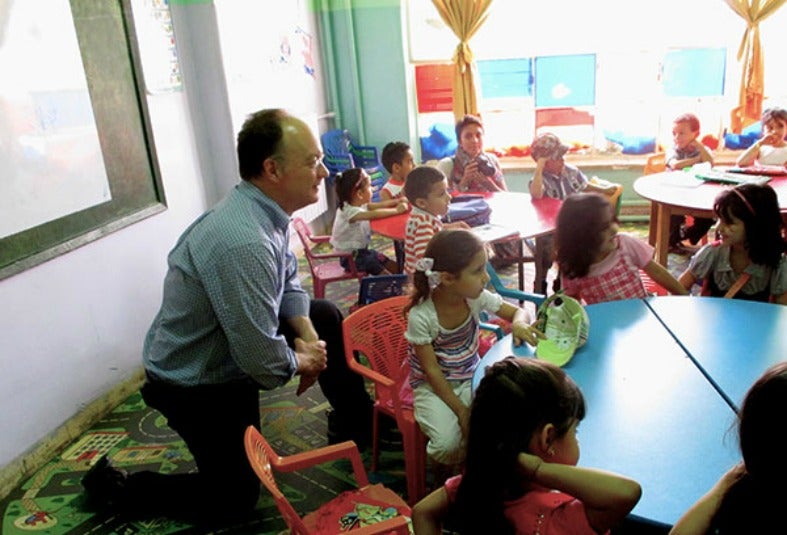 John J. DeGioia puts his leg up on a chair while listening to a speaker surrounding by children at a table