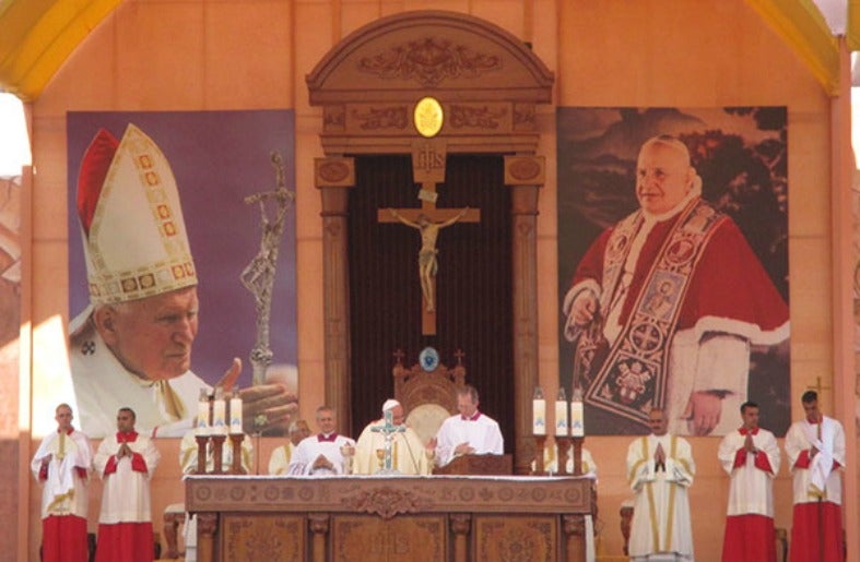 Pope Francis reads from a book on an alter with candles and servers to either side, with pictures of previous popes behind him