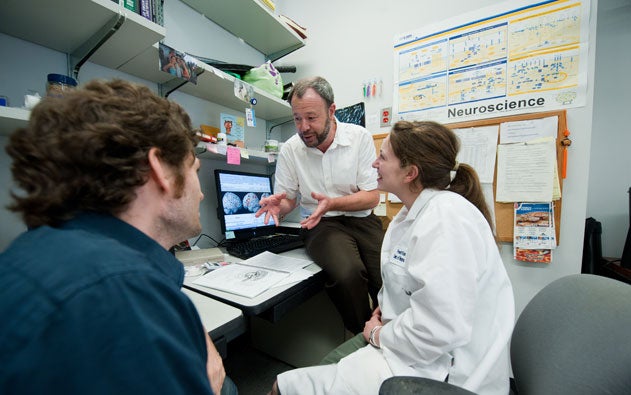 William Rebeck sits on a desk and talks to colleagues in an office. 