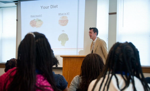 The backs of students heads are in the foreground as Bradley Bond speaks in a classroom near a podium with a projector screen