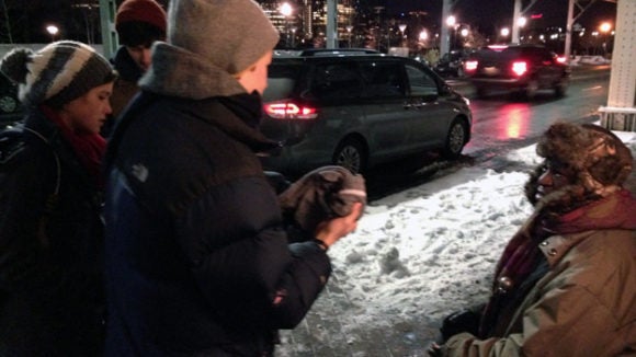 Students hand a jacket to a man sitting on a sidewalk on a snowy night