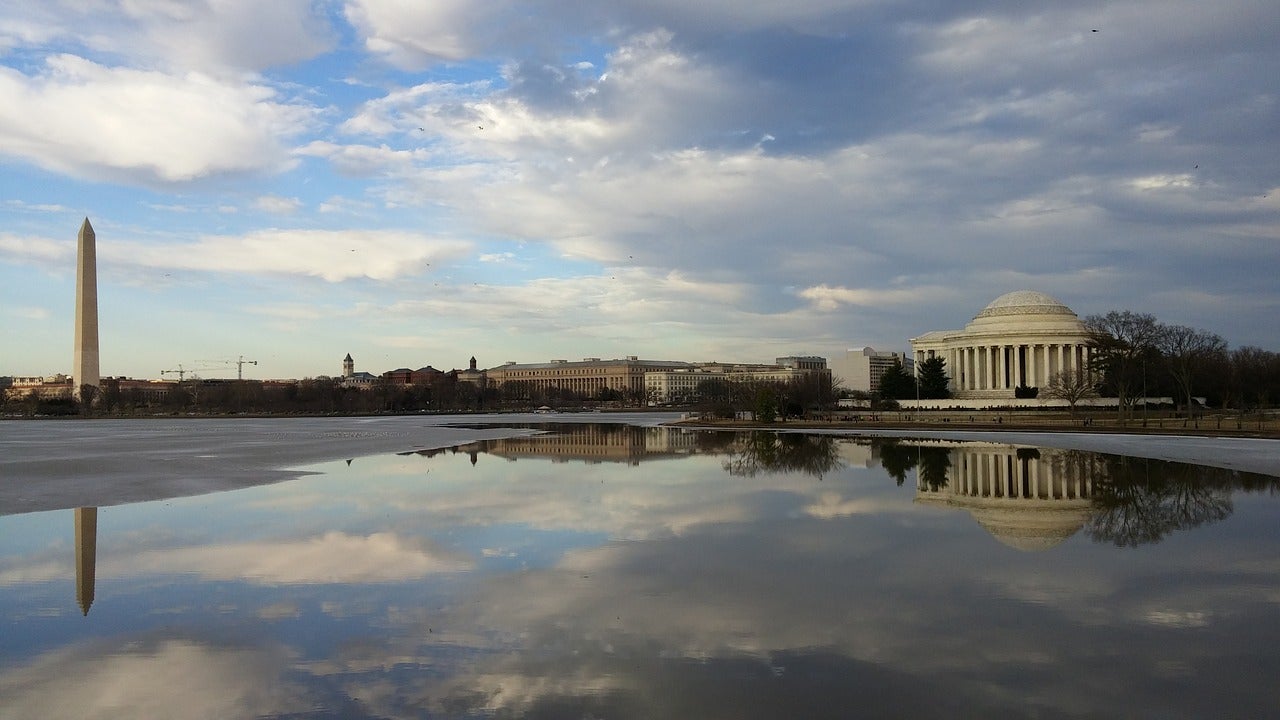 A wide shot of the sunny sky reflected in a pool of rain, the national monument and Jefferson memorial rise in the background