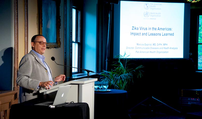 Marcos Espinal standing at podium with projector screen that reads "Zika Virus in the Americas: Impact and Lessons Learned"